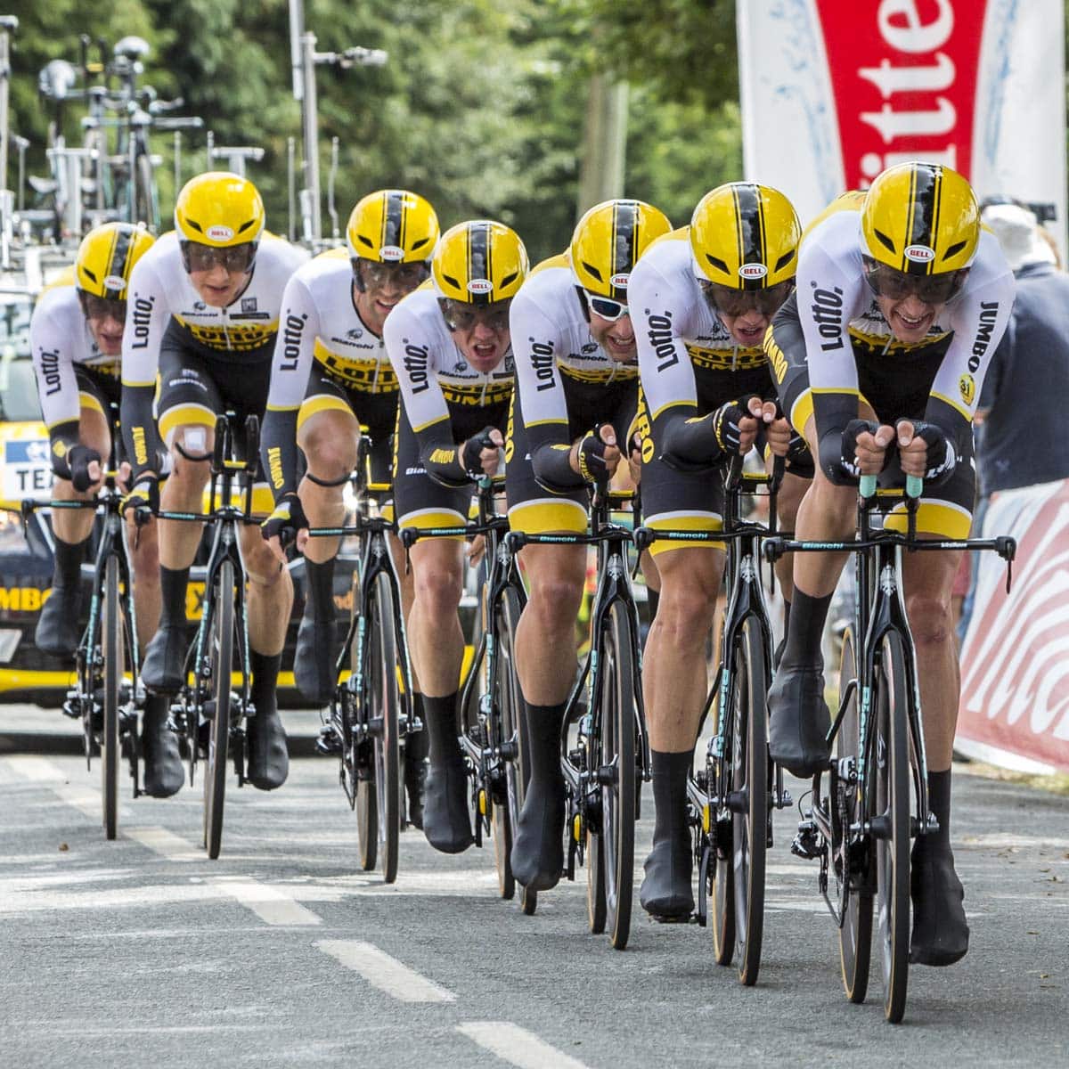 A professional cycling team in matching yellow and black helmets and gear rides closely together in a team time trial on a tree-lined road, with support cars following behind.