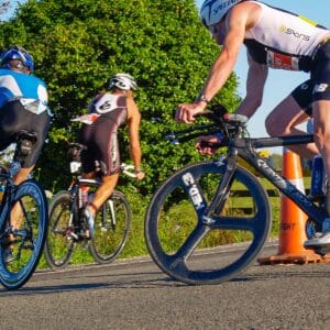 Two cyclists in a race navigate a sharp turnaround marked by an orange traffic cone. Both are wearing helmets and racing gear.