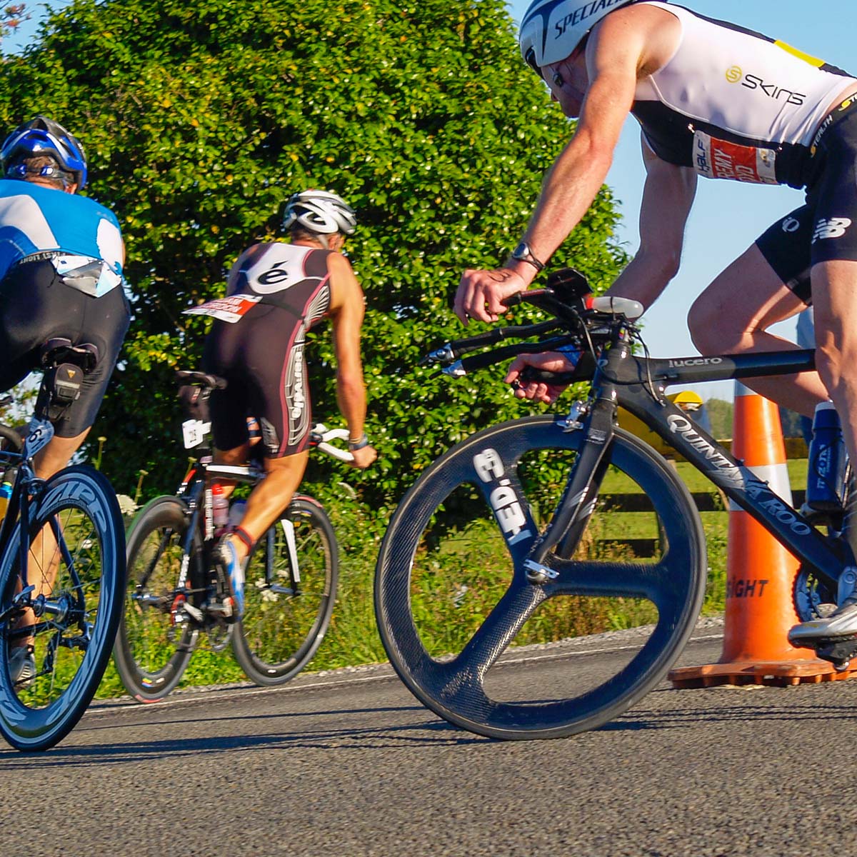 Two cyclists in a race navigate a sharp turnaround marked by an orange traffic cone. Both are wearing helmets and racing gear.