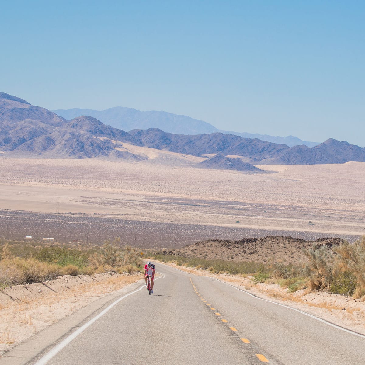 A lone cyclist rides on an empty road through a vast desert landscape during a Vegas In 24 ultra-cycling challenge, with dry shrubs in the foreground and rugged mountains in the distance under a clear blue sky.