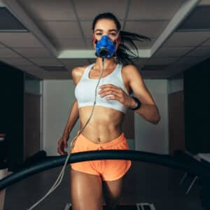 A woman wearing an athletic outfit and a blue oxygen mask runs on a treadmill in a gym, with the mask connected to a tube for a fitness or VO2 max test.