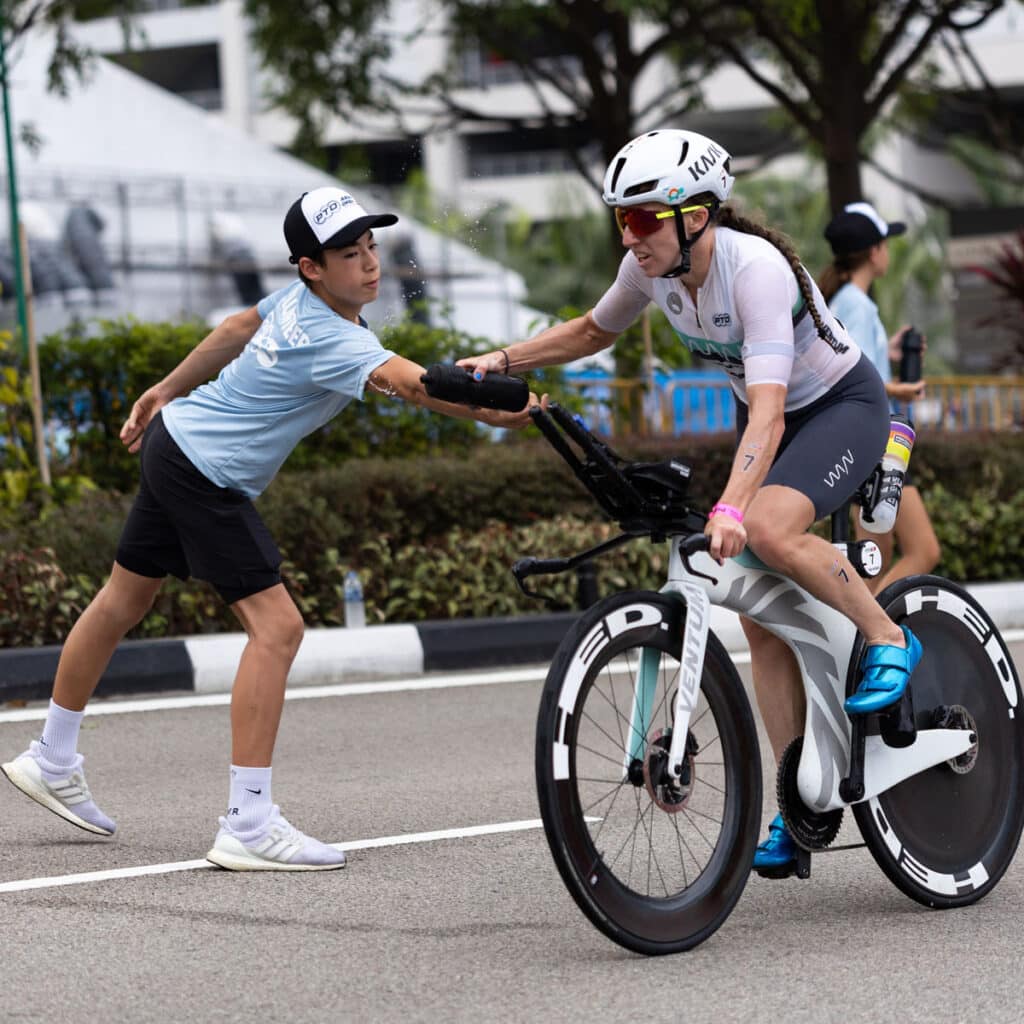 A cyclist in a race reaches for a water bottle handed by a volunteer on the roadside. The cyclist wears a helmet and sunglasses, while the volunteer is in a blue shirt and cap. Trees and buildings are visible in the background.