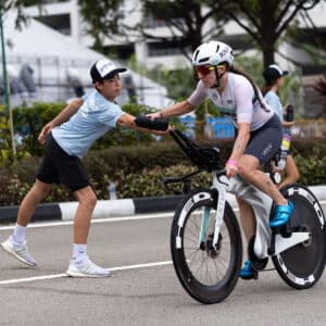 A cyclist in a race reaches for a water bottle handed by a volunteer on the roadside. The cyclist wears a helmet and sunglasses, while the volunteer is in a blue shirt and cap. Trees and buildings are visible in the background.
