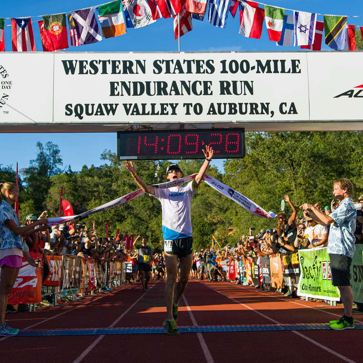 A runner crosses the finish line at the Western States 100-Mile Endurance Run, arms raised in victory, as spectators cheer beneath international flags.