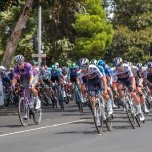 Cyclists in a competitive road race round a corner, closely packed together. Spectators and traffic lights are visible in the background. The image focuses on the concept of the 3 Kilometer Rule in cycling.
