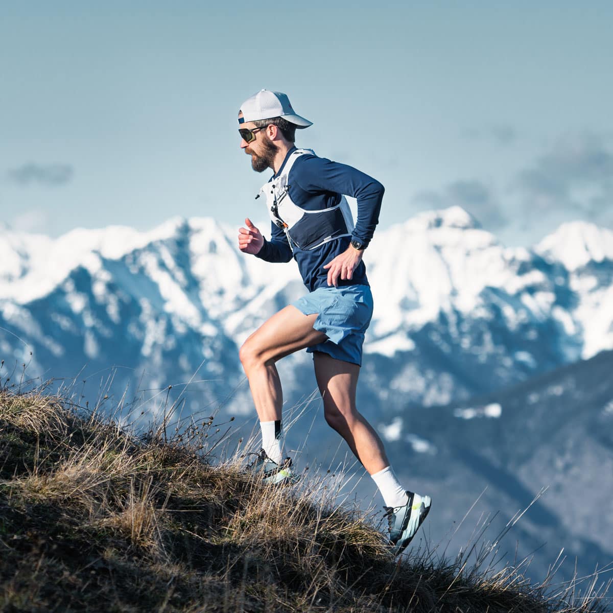 A man wearing a cap, sunglasses, and athletic gear runs uphill on a grassy trail, embracing altitude training with snow-capped mountains in the background under a clear sky.