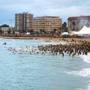 A large group of triathletes in swim gear runs from the sandy beach into the ocean, with tall buildings and white tents in the background. The event appears crowded and energetic, set on a lively city shoreline.