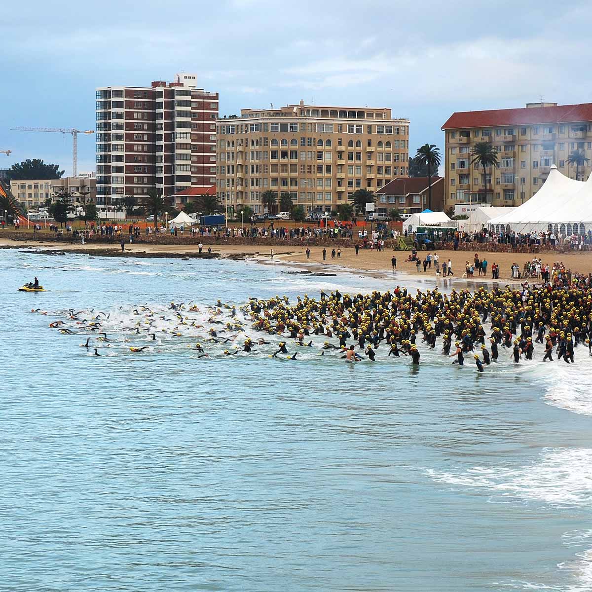 A large group of triathletes in swim gear runs from the sandy beach into the ocean, with tall buildings and white tents in the background. The event appears crowded and energetic, set on a lively city shoreline.