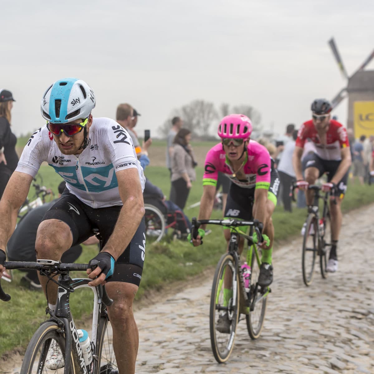 Three cyclists ride on a muddy cobblestone road during a race, with one rider in a white and blue kit leading. Spectators and a windmill are visible in the background.
