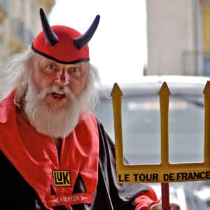 An older man with a white beard wears a red and black devil costume with black horns. He holds a yellow trident labeled Le Tour de France and stands outdoors, looking directly at the camera.