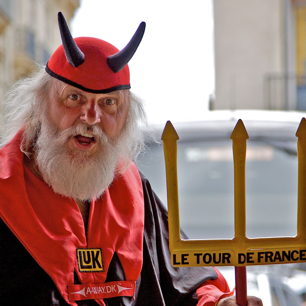 An older man with a white beard wears a red and black devil costume with black horns. He holds a yellow trident labeled Le Tour de France and stands outdoors, looking directly at the camera.
