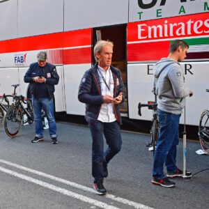 Three men stand near a team Emirates cycling bus; two check their phones while one walks past holding a phone and papers. Bicycles are parked nearby.