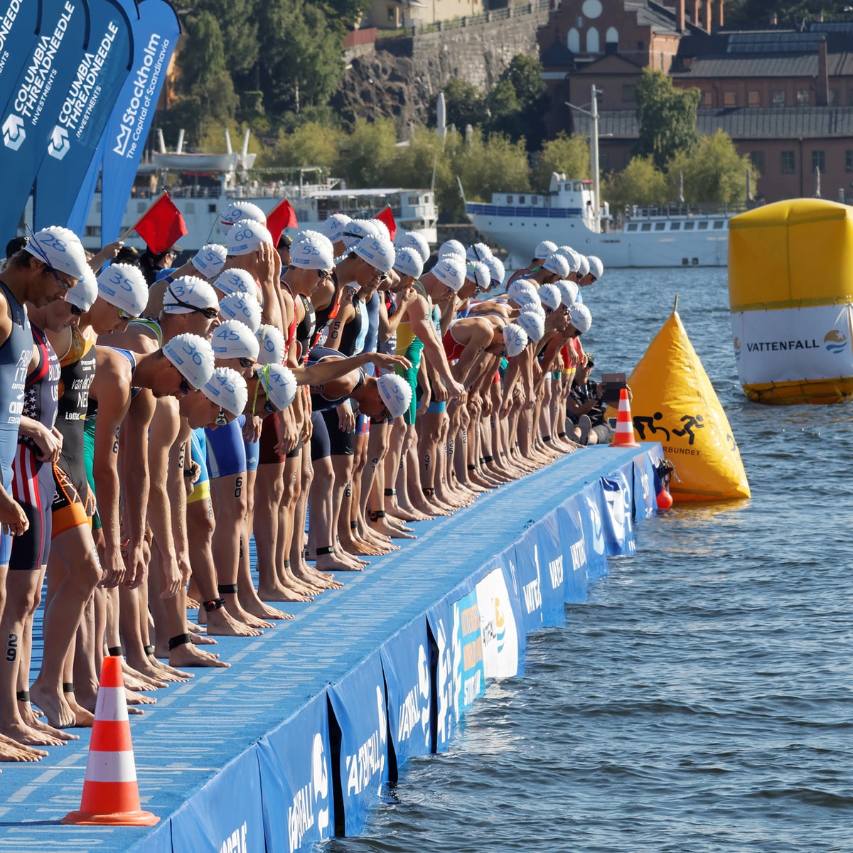 A group of triathletes in swim caps and wetsuits stand at the edge of a dock, preparing to dive into the water for a race. Boats and buildings provide a scenic backdrop.