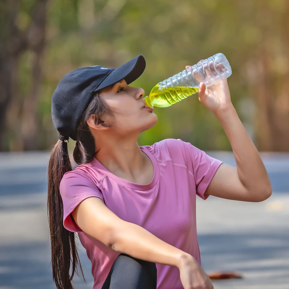 A woman in a pink shirt and black cap sits outdoors, drinking from a clear bottle with green electrolyte drink. The blurred greenery in the background suggests she is in a park or on a trail, replenishing her electrolytes after exercise.