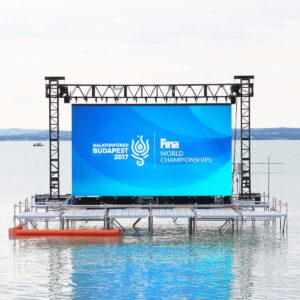 A large outdoor screen displaying Balatonfured Budapest 2017 FINA World Championships stands on a feeding pontoon above the water, with a calm lake and distant shoreline in the background.