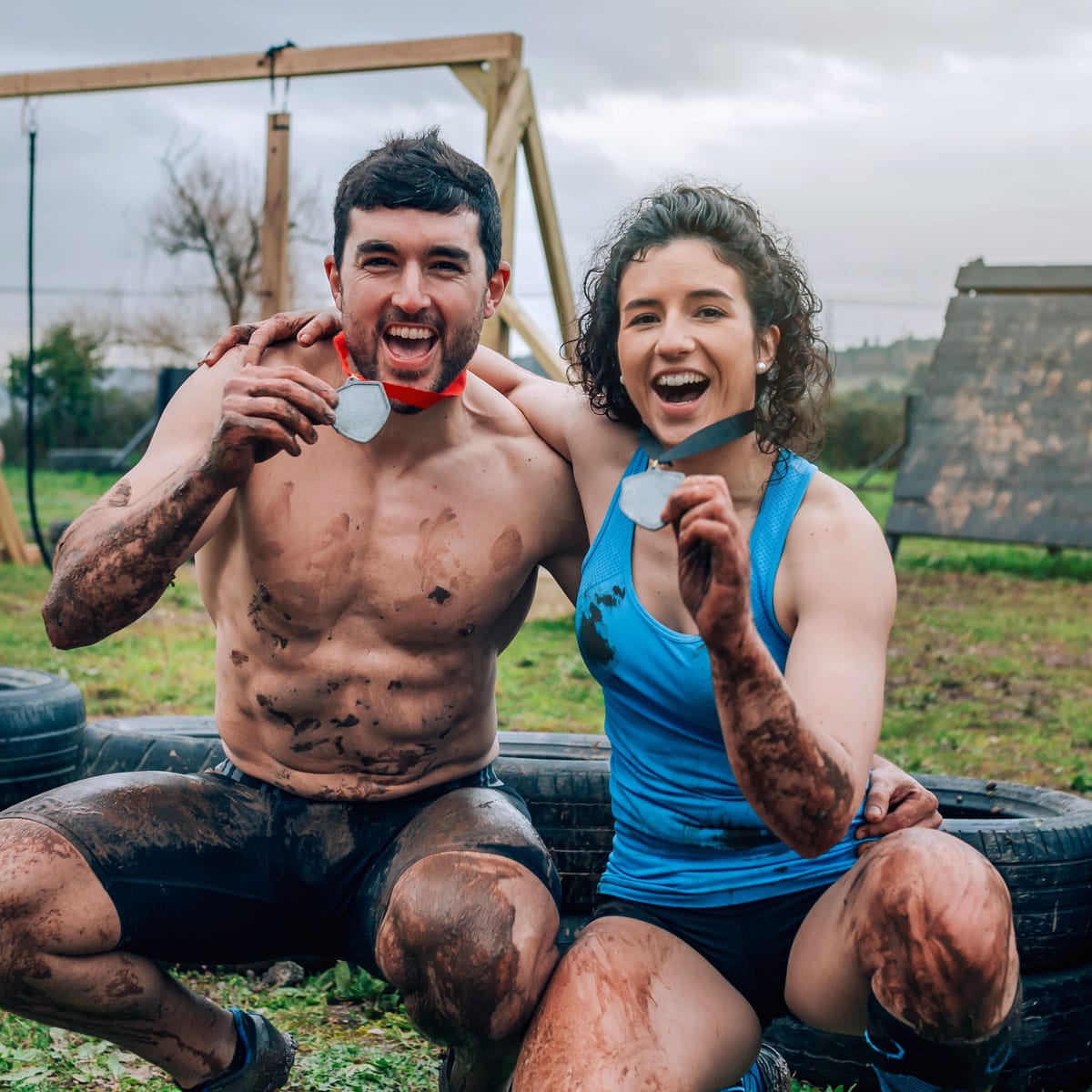 Two muddy runners, a shirtless man and a woman in a blue tank top, smile and hold up medals at an obstacle course event. The background features tires, wooden structures, and overcast skies.