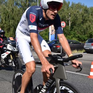 A male triathlete wearing a helmet and cycling gear rides a bike during a race, with a motorcycle and trees in the background.