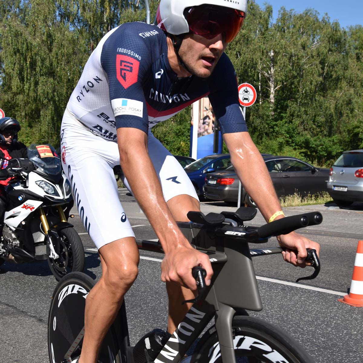 A male triathlete wearing a helmet and cycling gear rides a bike during a race, with a motorcycle and trees in the background.
