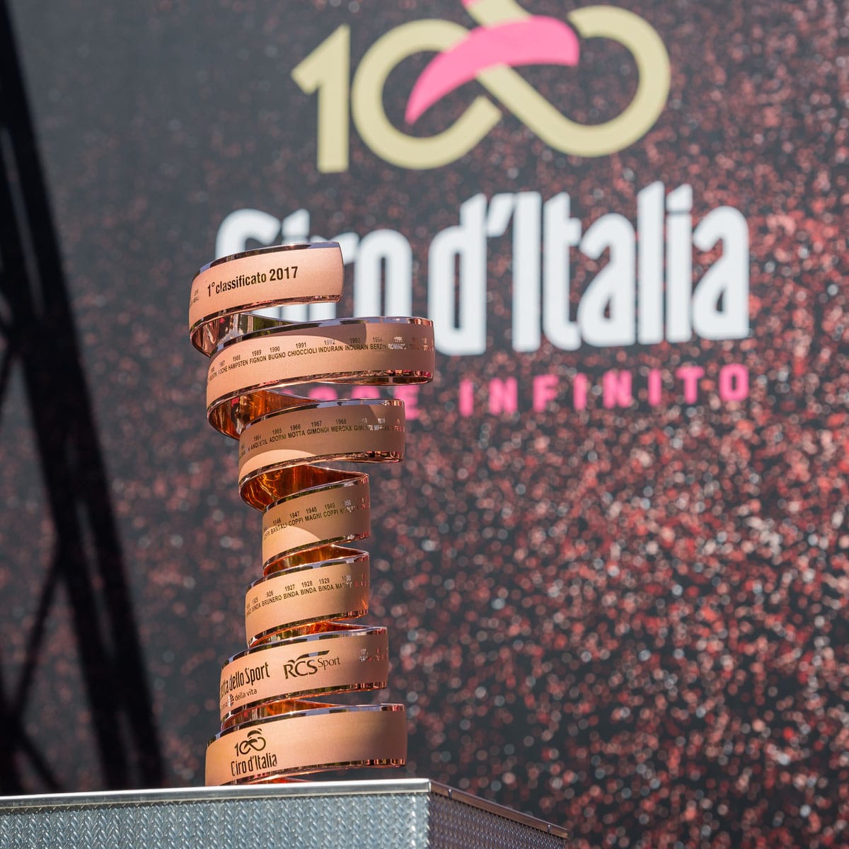 A close-up of the Giro d’Italia trophy, a spiraling golden sculpture with engraved text, is shown before a backdrop with the race’s logo and pink “100” design.