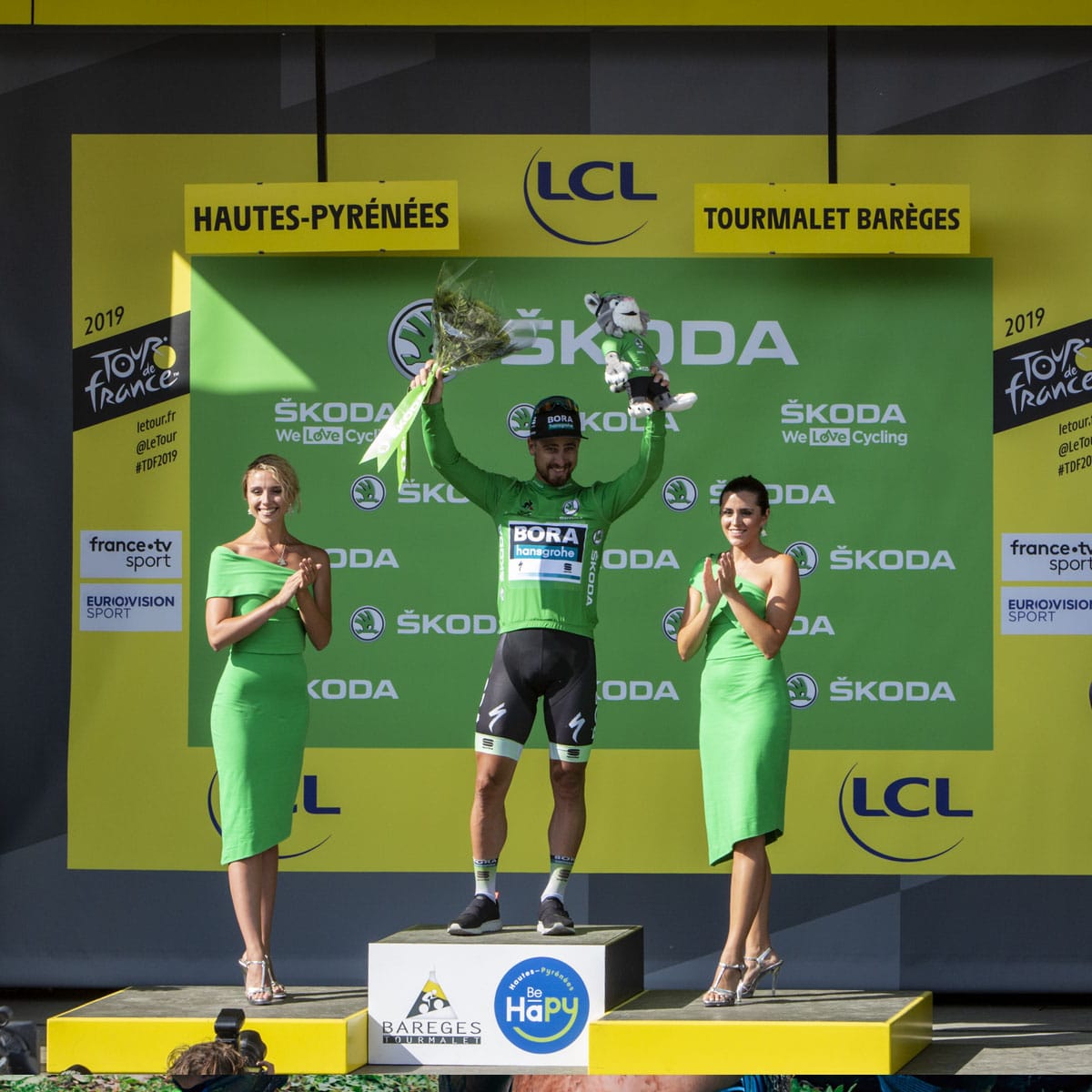 A cyclist in a green jersey celebrates on a podium, holding a trophy and flowers, flanked by two women in green dresses. The backdrop displays sponsor logos and yellow signage for the 2019 Tour de France.