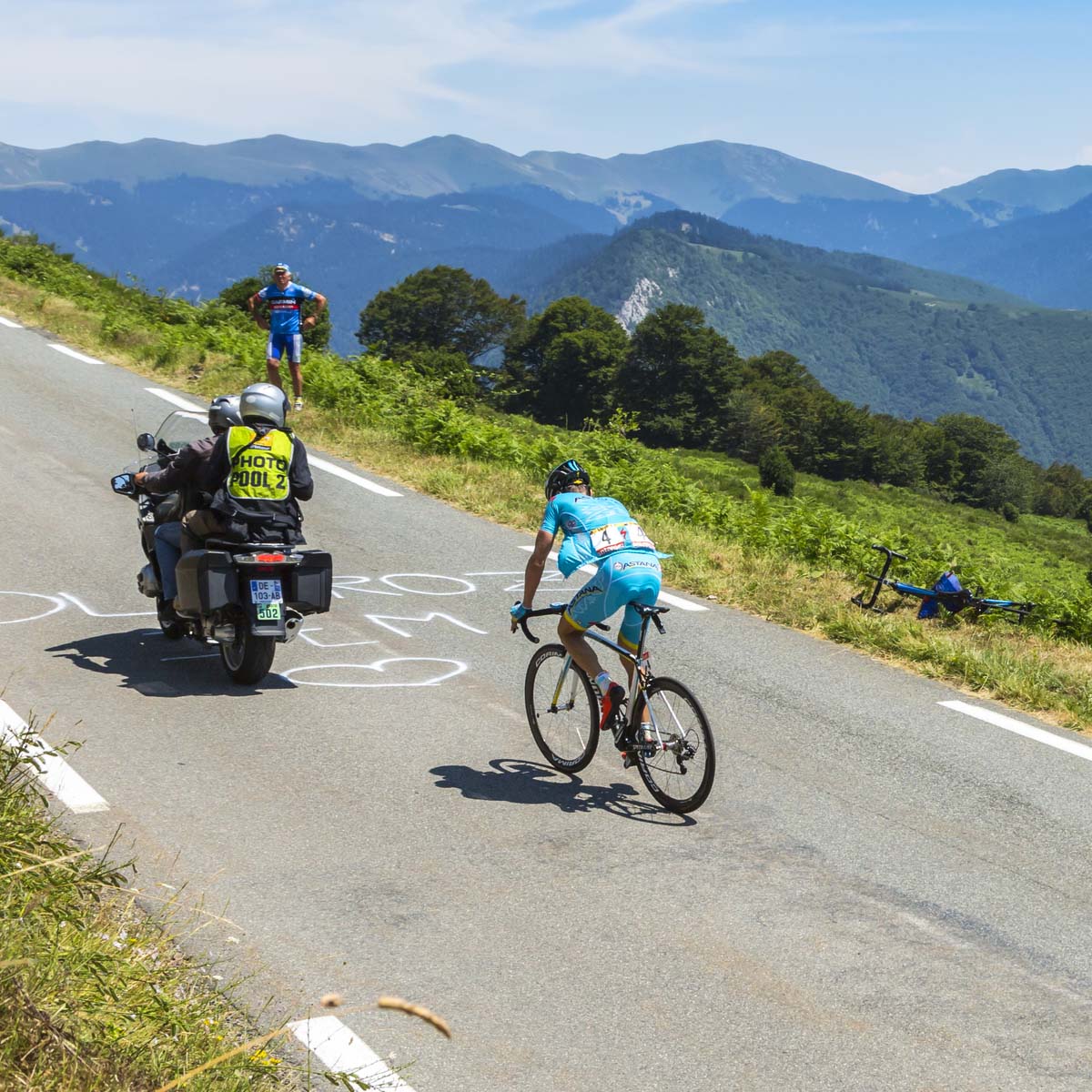 A cyclist climbs a winding mountain road, lead by a motorcycle with a cameraman, while two people cheer nearby. Green hills and mountains stretch under the blue sky.