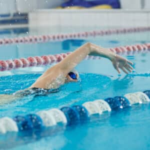 A swimmer in a blue swim cap and goggles performs a freestyle stroke in a pool, with one arm extended forward and water splashing beside them.