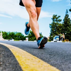 Close-up of a person running on a paved road with a yellow line, surrounded by trees and blue sky in the background. Other people and a parked car are visible in the distance.
