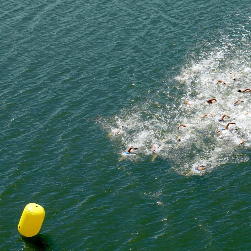 A group of swimmers competes in open water, creating splashes as they race toward an intermediate buoy floating in the greenish-blue water.