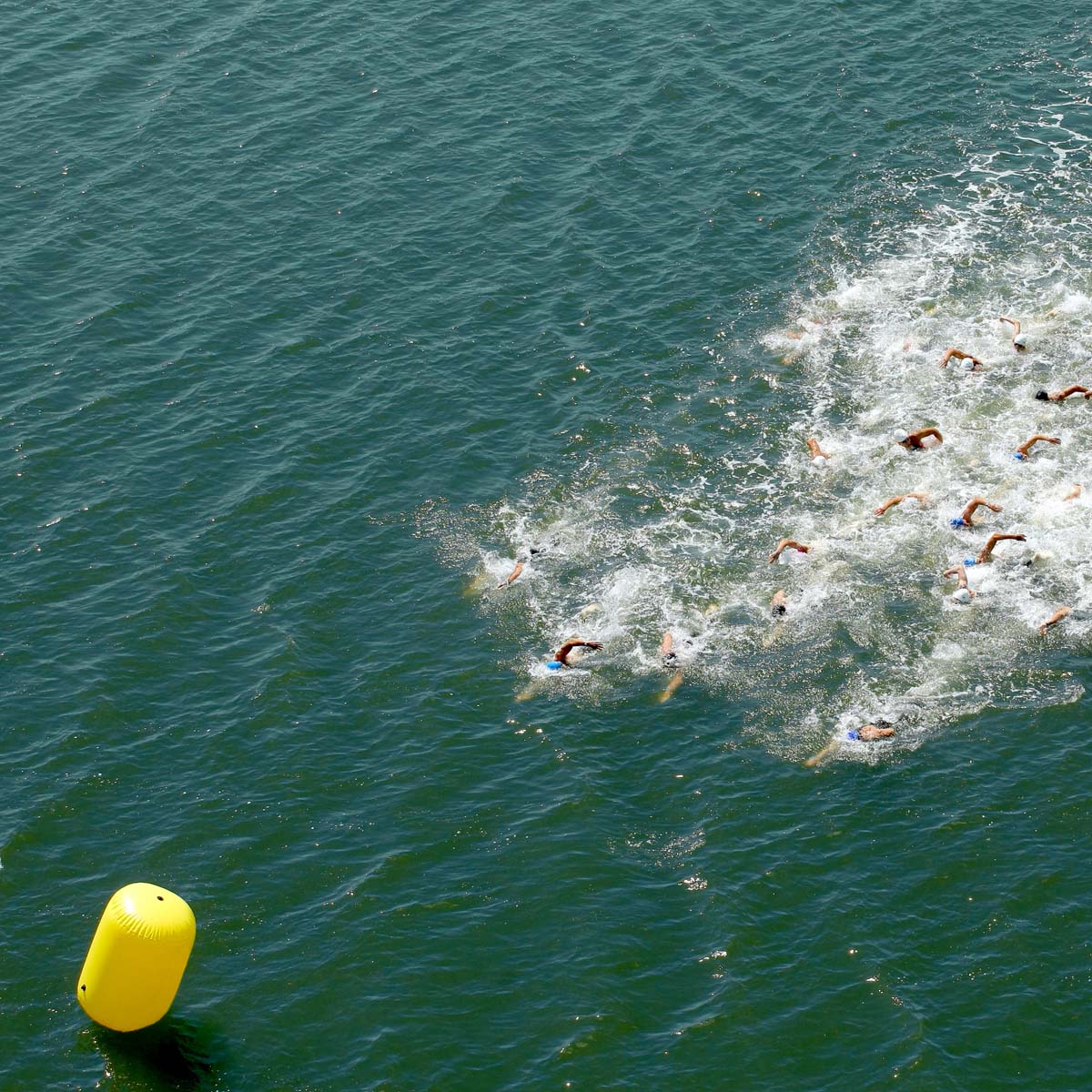 A group of swimmers competes in open water, creating splashes as they race toward an intermediate buoy floating in the greenish-blue water.