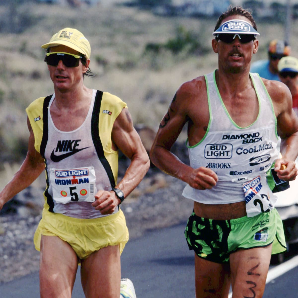 Two male triathletes run side by side outdoors during an Ironman event, wearing race bibs, sunglasses, and sports gear. The background shows a blurred, rocky landscape.
