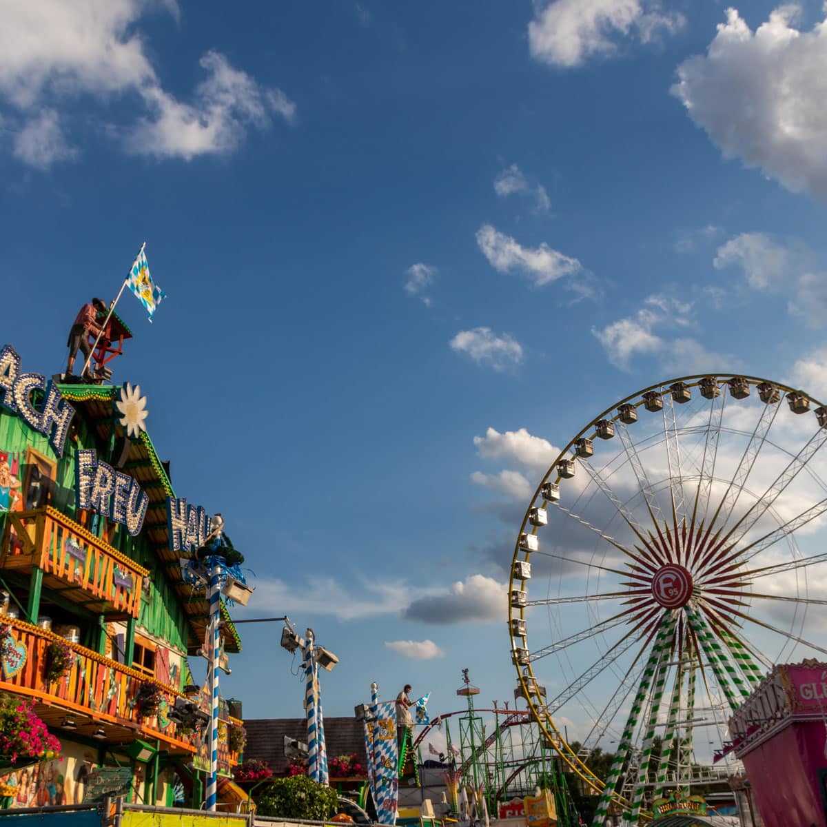 A colorful carnival scene with a large Ferris wheel on the right and a decorated building with balconies and flags on the left, all under a bright blue sky with scattered clouds.
