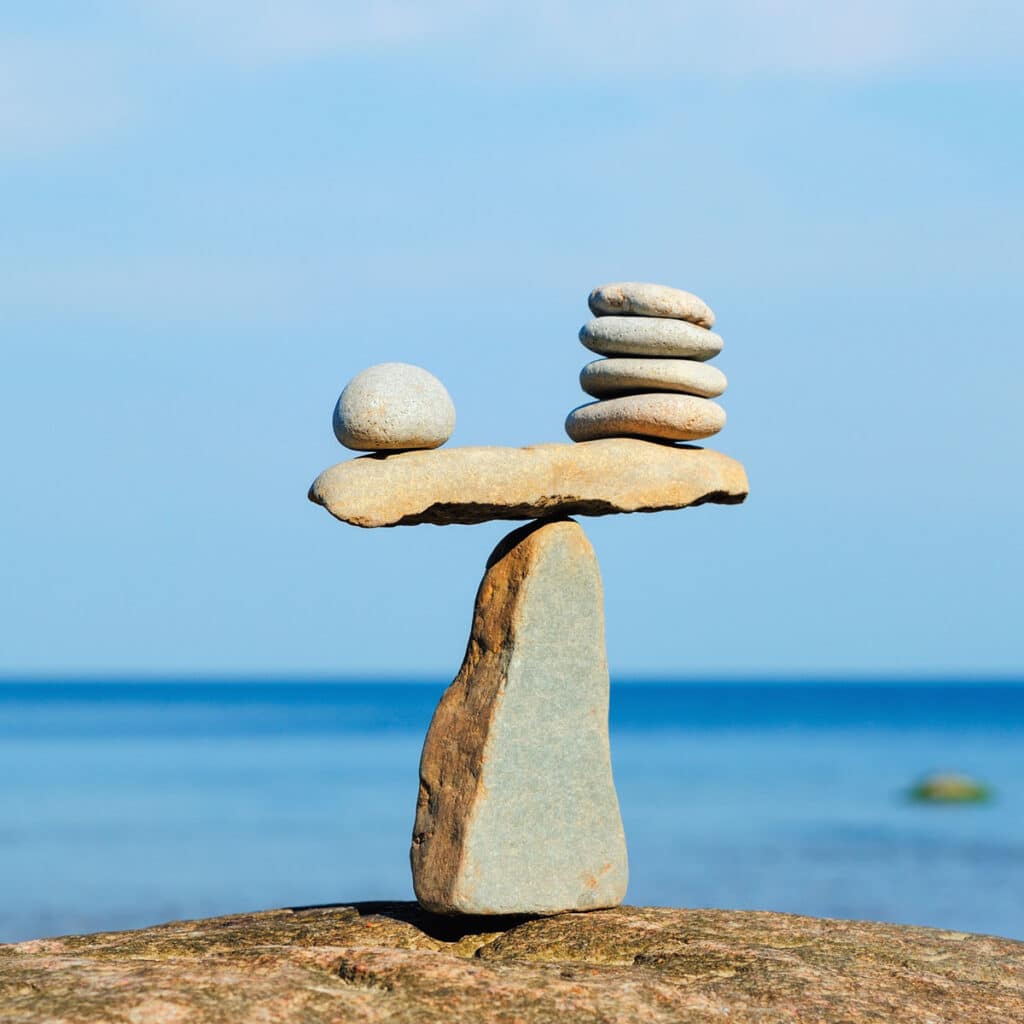 A balanced arrangement of stones achieves kinetic parity: one large upright rock supports a flat stone, with a round stone on the left and a stack of five flat stones on the right, set against a blue sky and sea background.