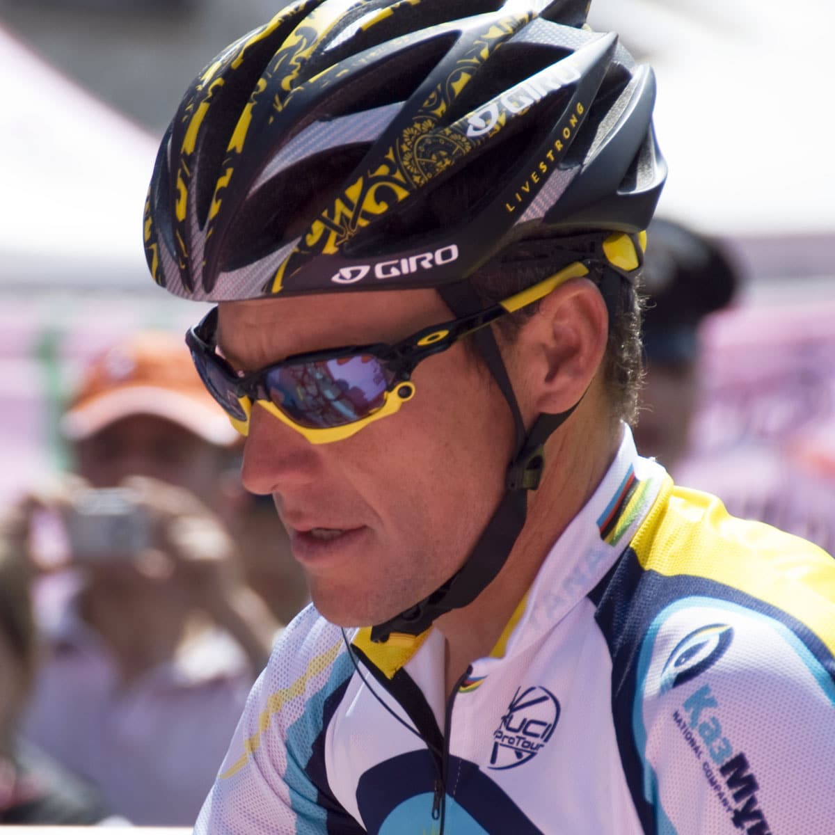 A cyclist wearing a black and yellow helmet, reflective sunglasses, and a white and blue cycling jersey with sponsor logos, participating in an outdoor event.
