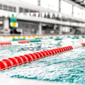 A close-up view of a swimming pool lane line divider with red floats, clear blue water, and a blurred background of the indoor pool area with lanes and overhead flags.