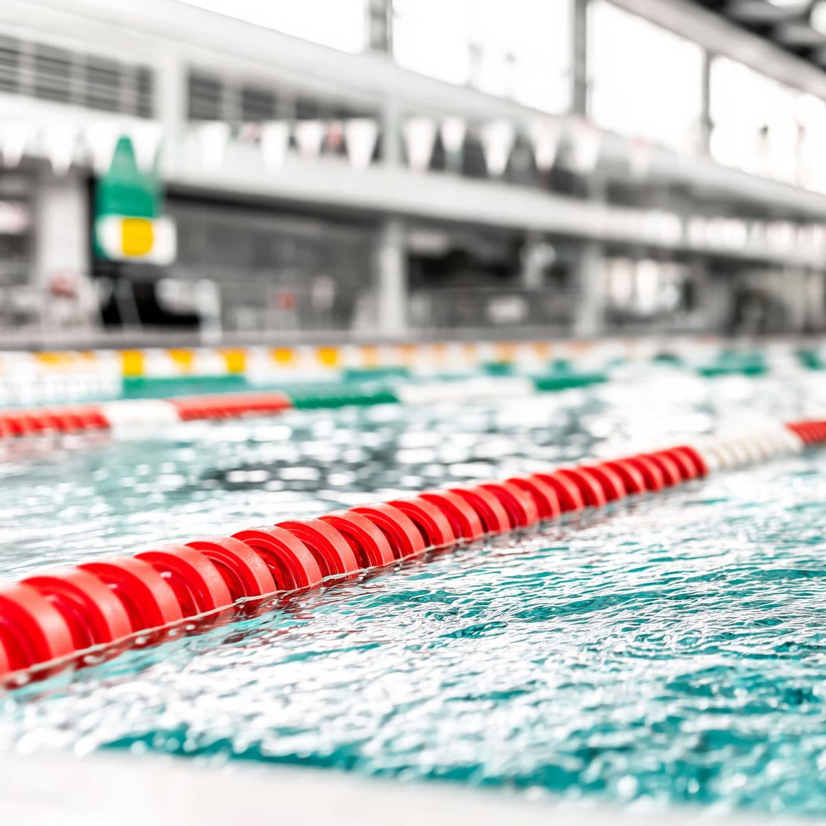A close-up view of a swimming pool lane line divider with red floats, clear blue water, and a blurred background of the indoor pool area with lanes and overhead flags.