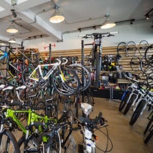 A local bike shop interior with various bicycles on display, including road and mountain bikes. Shelves on the wall hold accessories, parts, helmets, and wheels. The space is well-lit with ceiling lights.