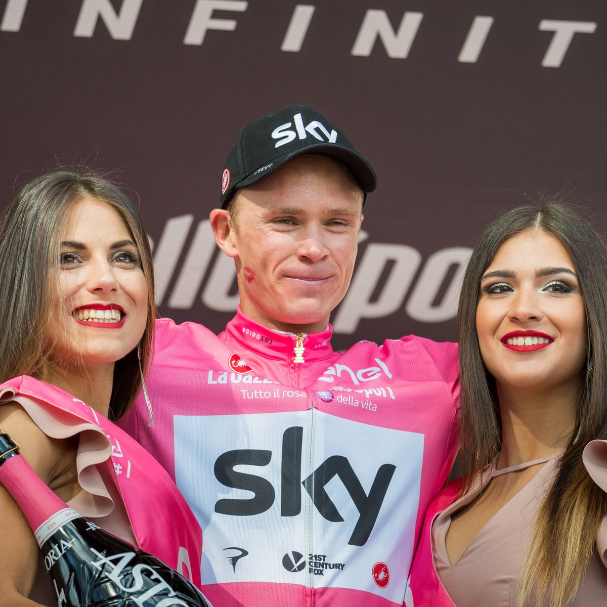 A cyclist in a pink jersey and black cap stands between two women holding a trophy bottle. All three are smiling in front of a branded backdrop.