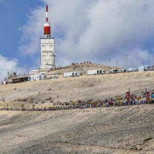 A white observatory building with a red-and-white antenna stands atop the rocky, barren summit of Mt. Ventoux—a legendary climb—with crowds, cyclists, and vehicles gathered along the winding road under a blue sky.