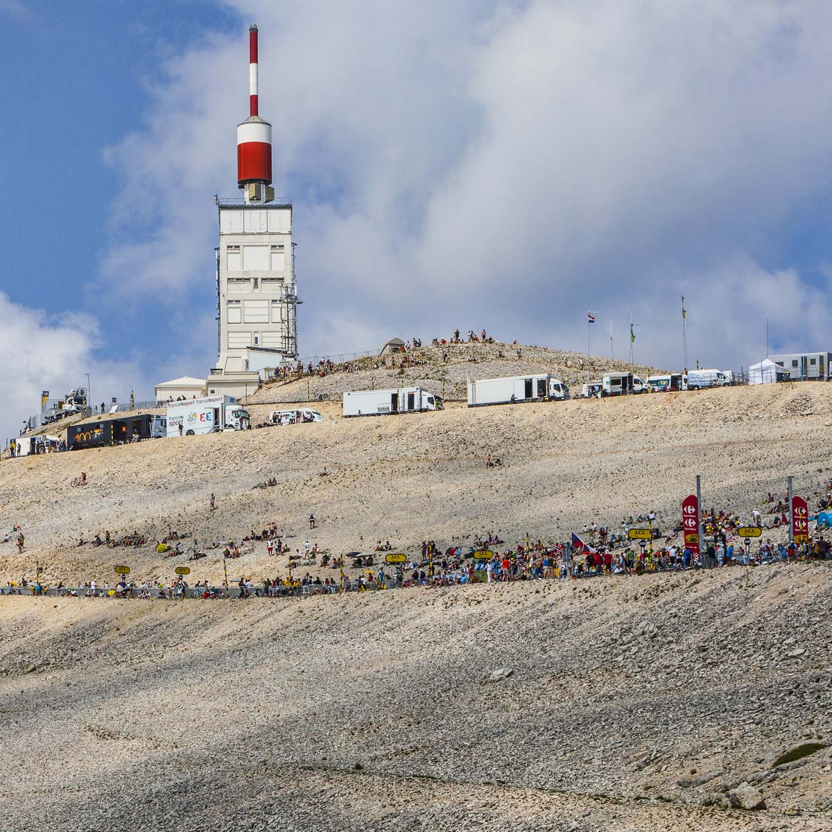 A white observatory building with a red-and-white antenna stands atop the rocky, barren summit of Mt. Ventoux—a legendary climb—with crowds, cyclists, and vehicles gathered along the winding road under a blue sky.