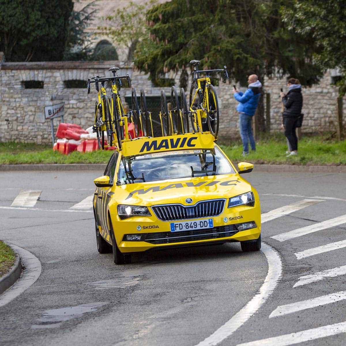 A yellow Mavic support car with bicycles on the roof drives along a road during a cycling event. Spectators in jackets stand nearby, while trees and a stone wall are visible in the background.
