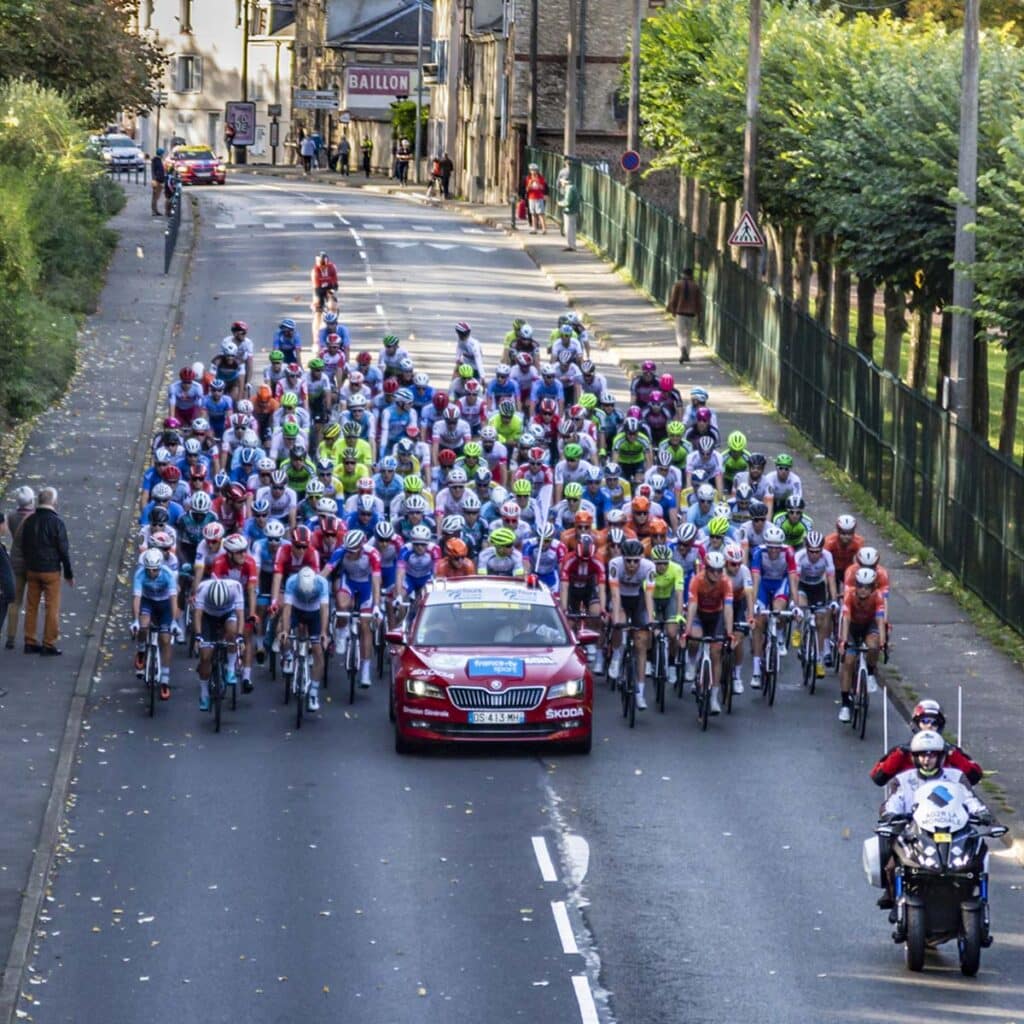 A large group of cyclists rides closely together behind a red car and motorcycle on a city street, marking the neutralized start of a race. Spectators watch from sidewalks, with buildings and greenery lining the road.