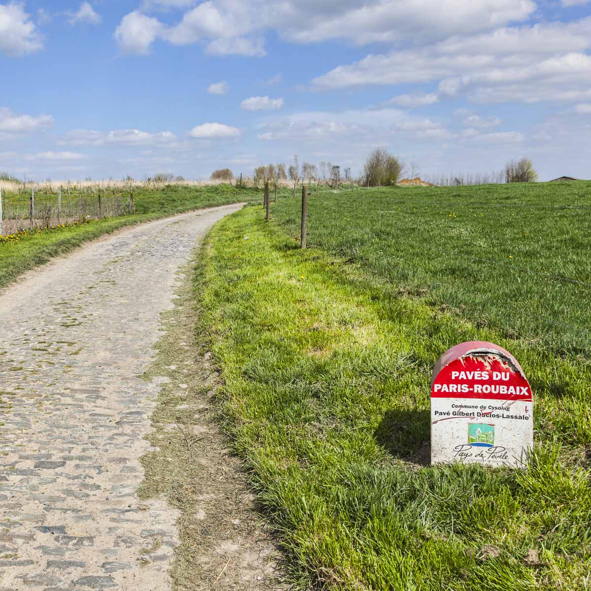 A cobblestone road winds through grassy fields beneath a blue, partly cloudy sky. A Paris-Roubaix race marker stands nearby.