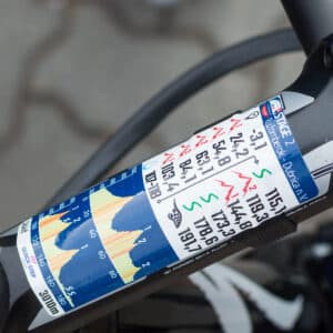 A close-up of a bicycle handlebar with a profile sticker displaying an elevation profile chart for a cycling race, showing various distances, heights, and climbs.