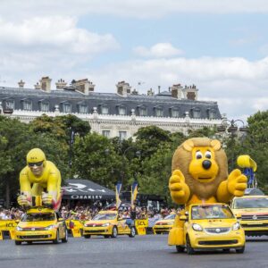 Colorful parade floats shaped like cyclists ride on yellow cars in front of a crowd and classic French buildings to promote the Tour de France during a festive pre-race event.