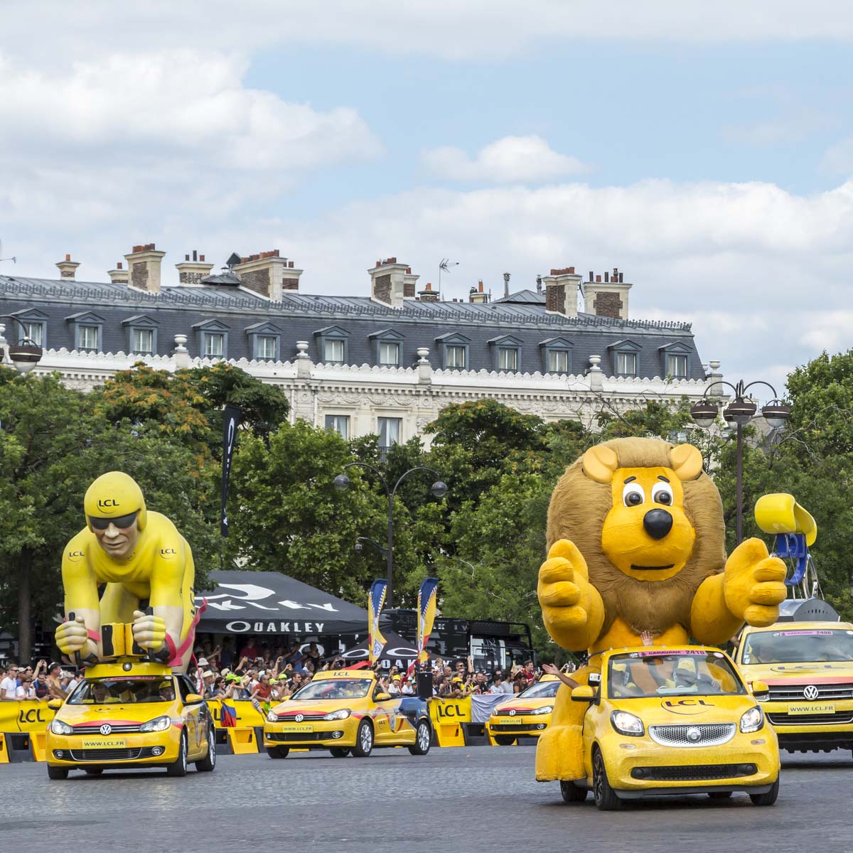 Colorful parade floats shaped like cyclists ride on yellow cars in front of a crowd and classic French buildings to promote the Tour de France during a festive pre-race event.