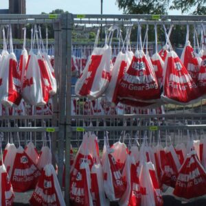 Rows of red-and-white Run Gear Bag bags hang on metal racks outdoors in a triathlon T2 transition zone, with a large church and trees in the background.