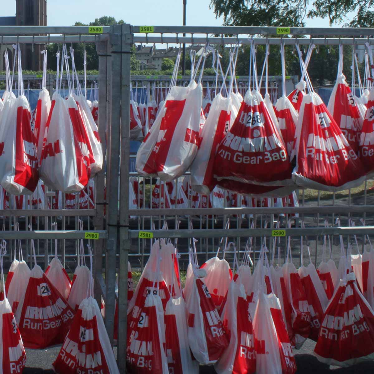 Rows of red-and-white Run Gear Bag bags hang on metal racks outdoors in a triathlon T2 transition zone, with a large church and trees in the background.