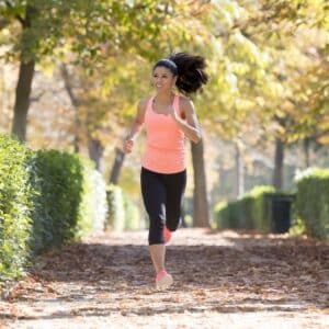 A woman in athletic wear jogs along a sunlit, tree-lined path covered with fallen leaves, smiling.