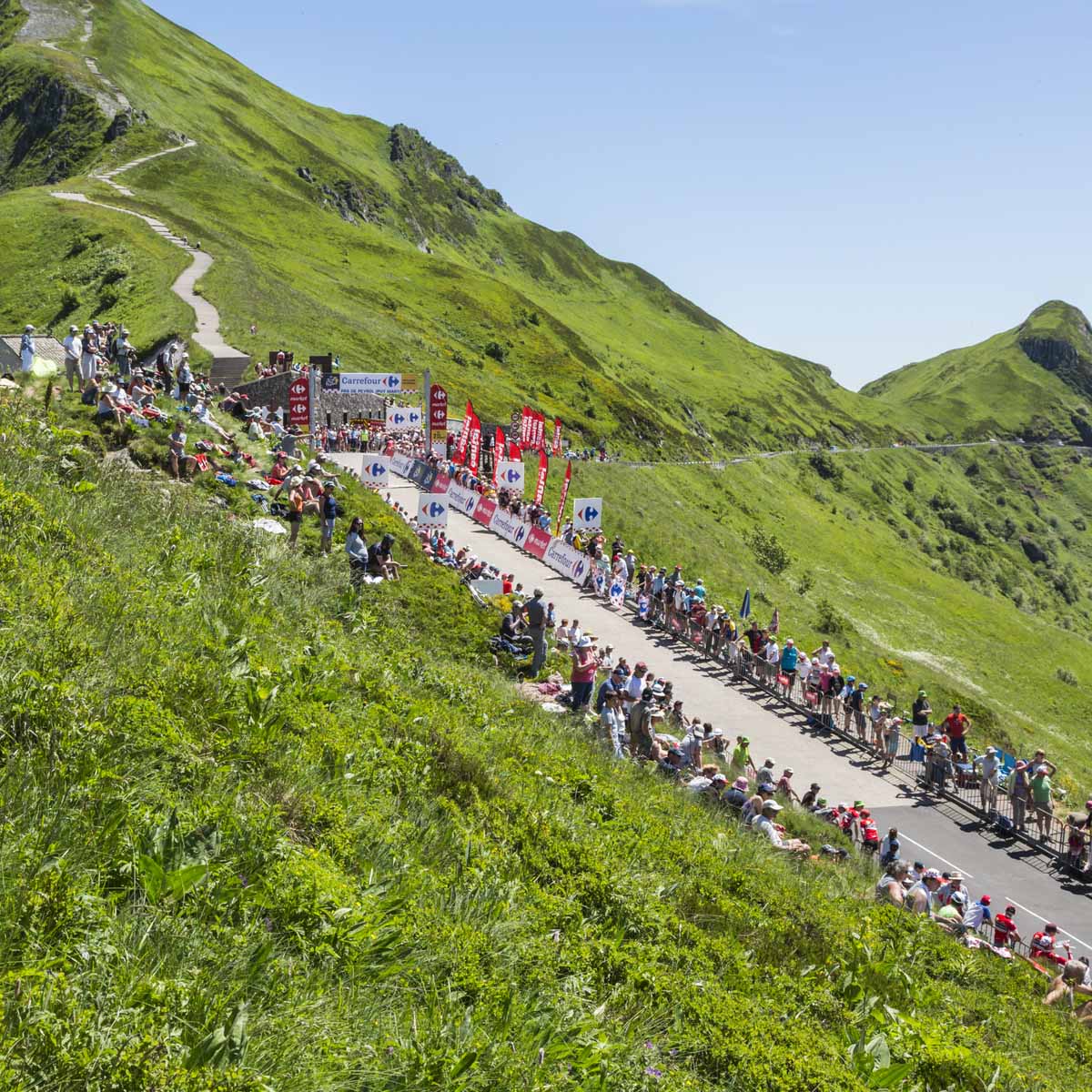 A winding mountain road lined with spectators and banners hosts a summit finish cycling event, surrounded by lush green hills under a sunny sky.