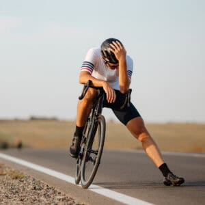 A cyclist wearing a helmet and sport clothes sits slumped on his bike at the roadside, head in hand, looking exhausted or distressed on an empty rural road.